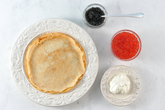 Pancakes thin on a large white plate. There are two cups with red and black caviar. There is sour cream on the saucer. Light background. Close-up. View from above.