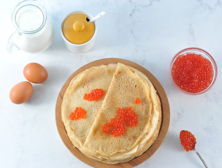 Pancakes thin on a round wooden tray. On the pancakes laid red caviar. Next to a bowl of red caviar.In the frame sugar bowl, a jug of milk and chicken eggs. Close-up. View from above.