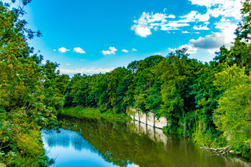 Fototapeta premium Fluss Kanal mit Bäumen in sattem grün am Ufer sonniges Wetter blauer Himmel Sommer