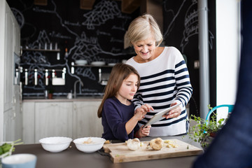 A small girl and grandmother with tablet cooking at home.