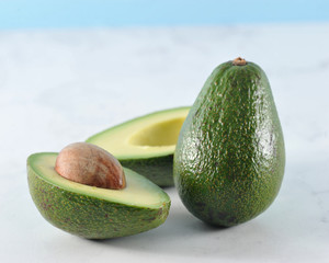 The avocado is green. One whole fruit, and the second cut in half. Pulp and large bone. Light marble background. Close-up. Macro photography.