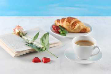 A cup of coffee and a crispy croissant. There are two candies in the form of heart. In the frame an open book, on the pages of the book lies a flower. Light background. Close-up.
