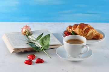A cup of coffee and a crispy croissant. Around the candy in the shape of the heart. In the frame an open book, on the pages of the book lies a flower. Light background. Close-up.