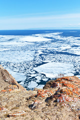 Time of spring ice drift on Baikal Lake. Top view of coastal rocks with red lichen and white ice on blue water