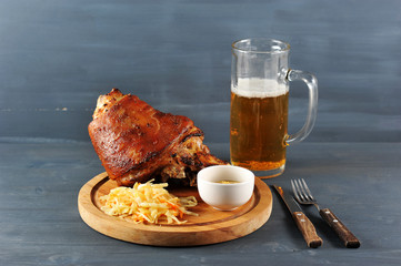 Pork shank on a wooden tray. Next stewed cabbage and a cup of mustard. In the frame a glass of light beer and cutlery. Dark wooden background. Close-up.