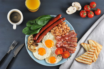 English breakfast - brunch. On a plate fried bacon, sausages, fried fresh tomatoes and champignons, white beans in tomato sauce, toast, fried eggs. Close to orange juice and coffee. View from above.