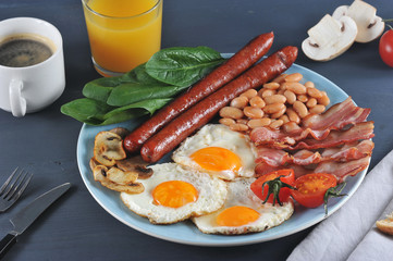 A classic English breakfast of seven ingredients. Fried bacon, sausages, fried fresh tomatoes and champignons, white beans in tomato sauce, toast, fried eggs. Dark background. Close-up.