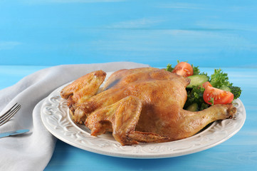 On a white plate, a chicken cooked on a grill. Next to a salad of fresh tomato, cucumber, lettuce leaves. In the frame napkin and cutlery. Blue wood background. Close-up.