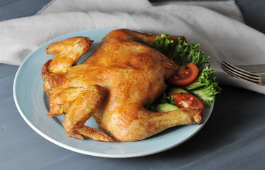 Grilled chicken. On a blue plate next to a chicken salad of fresh tomato, cucumber, lettuce leaves. In the frame napkin and cutlery. Dark wood background. Close-up. 