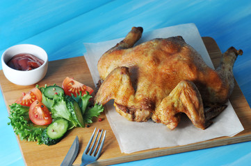 Chicken with golden and crispy crust. The chicken is cooked on the grill. On a wooden tray next to the chicken, fresh vegetables, a cup of ketchup and cutlery. Blue background. Close-up. 