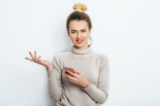 Horizontal portrait of displeased woman has indignant expression while holding smartphone, frowns eyebrows, can`t understand something, isolated on white background. Discontent emotion on face girl