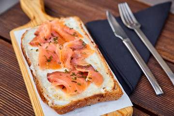 A sandwich with melted cheese and slices of salmon on a light cutting board and a brown wooden table. Photographed with natural light. Metal fork and knife on a black napkin.