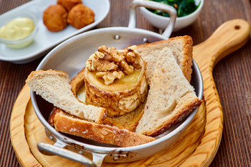 Snack from oven-baked goat cheese with walnuts and roasted homemade bread in an aluminum saucepan on a round light wooden cutting board on a brown wooden table. With cheese balls and sauce.