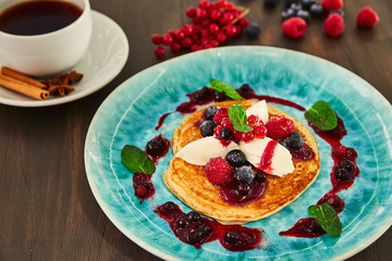 A large homemade pancake with white mascarpone cream, raspberries, blueberries, currants, berries and honey on a blue plate. It is decorated with jam from mountain ash and green young leaves of mint.