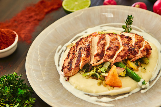 Grilled chicken fillet is cut into slices on a cushion of mashed potatoes with vegetables - onion, garlic, pepper, cabbage, radish, greens in a large beige dish on a dark wooden table.