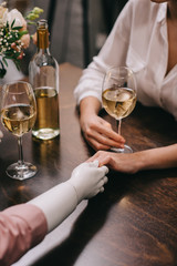 cropped shot of woman and mannequin holding hands at table with glasses of wine, unrequited love concept