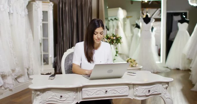 4K Young woman working on laptop in bridal wear boutique, with elegant wedding gowns on display in background. Slow motion.