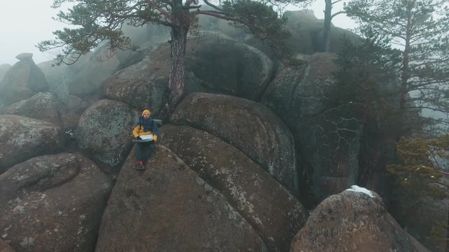 Air view at the man sitting on the top of the rock in winter mountains covered with fog