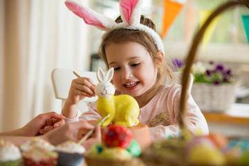 A mother and daughter celebrating Easter, painting bunny with brush in yellow color. Happy family smiling and laughing. Cute little girl in bunny ears preparing the holiday.