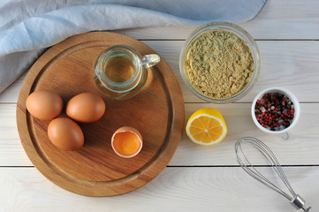 Set for the preparation of mayonnaise. On a wooden tray, a jug with olive oil and chicken eggs. Next to half a lemon, a bowl of mustard powder,  spices. Light background. Close-up. View from above.