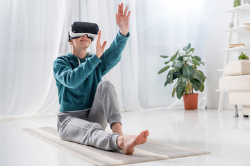 girl sitting on yoga mat with virtual reality headset and touching something at home