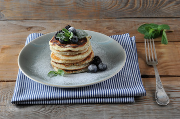 Appetizing pancakes are decorated with blueberries, blackberries, mint and sprinkled with powdered sugar. Below the plate is a napkin, next to the fork. Wooden  background. Close-up.