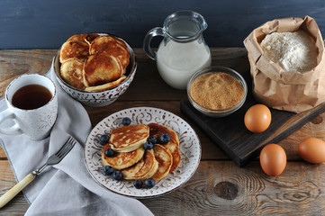 Plate with pancakes and blueberries. Next to the bowl with pancakes, sour cream and a cup of tea. In the frame, products for the preparation of pancakes: flour, eggs, sugar, milk. Wooden background. 