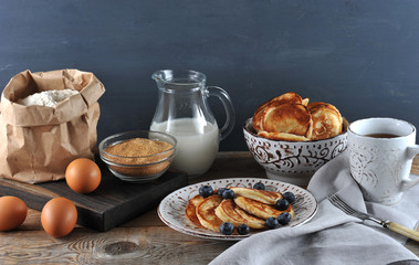 Plate with pancakes and blueberries. Next to the bowl with pancakes, sour cream and a cup of tea. In the frame, products for the preparation of pancakes: flour, eggs, sugar, milk. Wooden background. 