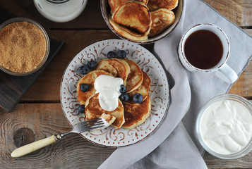 Pancakes with blueberries and sour cream. Next fork. In the frame, a bowl of pancakes, cane sugar, sour cream and a cup of tea. Wooden background. View from above.