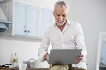Being glad. Happy mature man keeping smile on his face and holding laptop while reading message