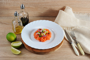 Tartar from salmon. The dish is decorated with pieces of avocado, capers, red onions. In the frame, lime, napkin, cutlery, containers with butter and balsamic sauce. Light wooden background. Close-up.
