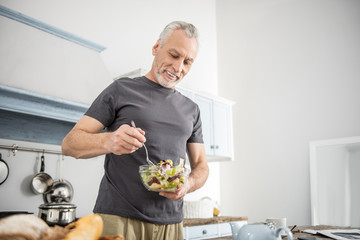 Eat vitamins. Cheerful pensioner being in the kitchen and keeping smile on his face while mixing salad