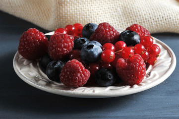 Juicy and ripe berries of raspberries, blueberries and red currants in a wooden bowl. The berries are indicated by a leaf of mint. In the frame cloth napkin. Dark background. Close-up. View from above