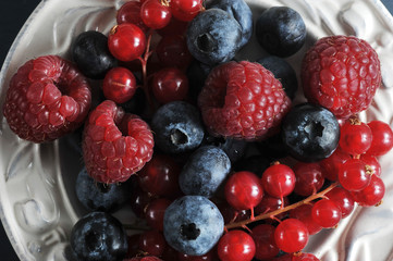 Juicy and ripe berries of raspberries, blueberries and red currants on a white plate. Dark background. View from above. Close-up. Macro photography.