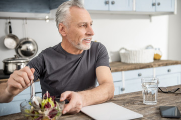 Dinner preparation. Happy bearded male person keeping smile on his face and turning head while mixing salad