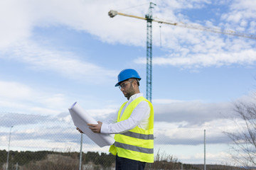 Architect or Engineer man holding blueprints on the Construction Site. Wearing protection clothes. Crane on background over blue sky. Job concept. Back light