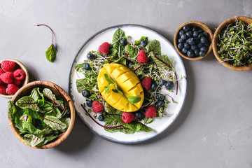 Green vegan salad with berries strawberry, blueberry, sprouts, young beetroot leaves, mango on plate. Ingredients in wooden bowls above over grey concrete background. Top view, space. Healthy eating