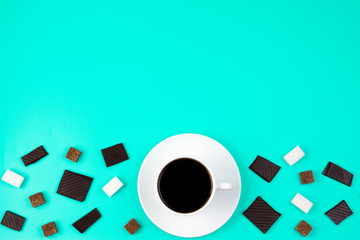 White cup of coffee, brown and white sugar cubes and chocolate pieces on turquoise background