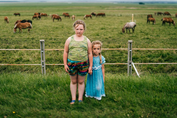 Obraz premium Tilt shift blurred expressive mood potrtrait of two little sisters resting outdoor in summer together. Young girls with emotional faces. Chilhood in village. Funny kids near grazing horses field.