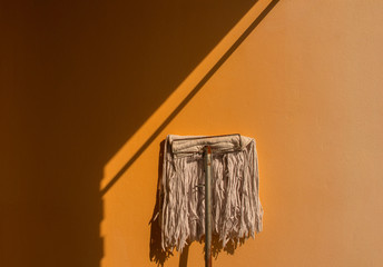 Old mop leaning against the orange wall have plenty of sunshine and shades.