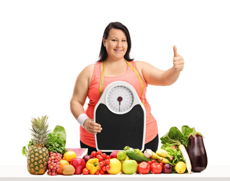 Overweight woman with a weight scale making a thumb up gesture behind a table with fruit and vegetables