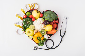 vegetables and fruits laying in heart shaped dish near stethoscope and measuring tape isolated on white background