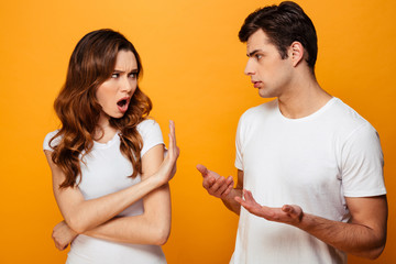 Photo of angry man and woman in white t-shirts being in fight while posing on camera with stop gesture, isolated over yellow background