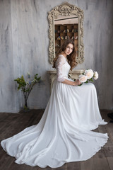 Beautiful bride sitting with flowers near the mirror. Floor-length dress. Vintage mirror. Large pink roses. Gray wall.