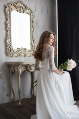 Beautiful bride sitting with flowers near the mirror. Open back. Floor-length dress. Lace. Vintage mirror. Boudoir. Large pink roses. Gray wall.