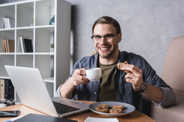 young handsome man working with laptop and eating cookies and coffee
