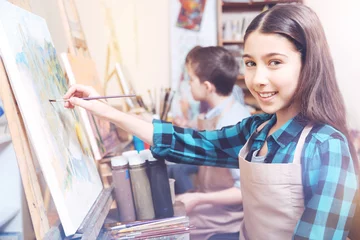 Fotobehang Schilderij My top hobby. Beautiful dark long haired girl posing and smiling for the camera while creating her new masterpiece with oil paints.  © Viacheslav Yakobchuk