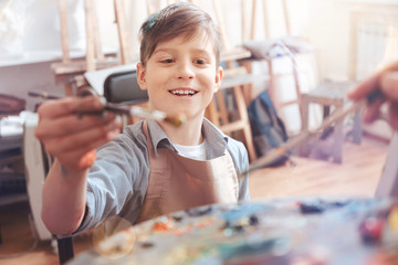 Never getting bored. Overwhelmed teenager grinning broadly while holding a painting brush and mixing oil paint while searching for a perfect.