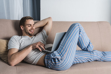 young relaxed freelancer in pajamas working with laptop on couch