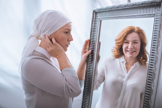 Upset Sick Mature Woman In Kerchief Looking At Smiling Reflection In Mirror, Cancer Concept